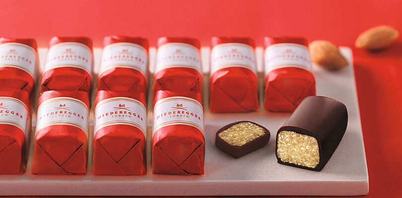 Marzipan Klassiker der Firma Niederegger in zwei Reihen aufgereiht, eingepackt in rote Wickler mit einer weißen Banderole. Auf der rechten Seite ist ein Klassiker angeschnitten worden.